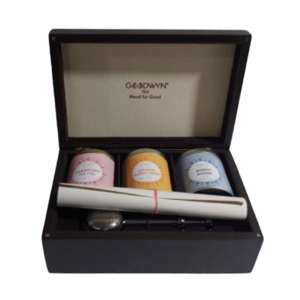 A wooden gift box containing three tins of tea, a rolled-up paper tied with a red string, a tea infuser, and a small spoon. The inside lid is labeled “GOODWYN Tea Hand for Good.”.